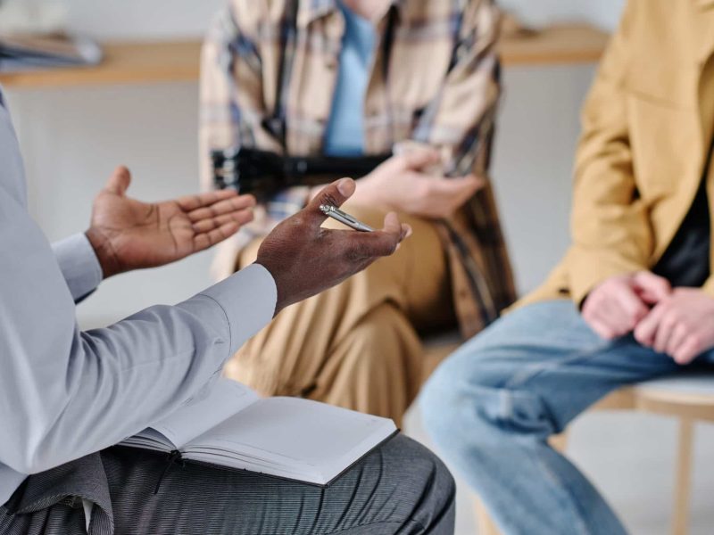 Gespreksgroep tijdens een therapiesessie in een afkickkliniek, met close-up van gebarende handen en een notitieboek.
